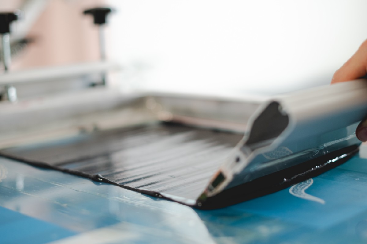 Selective focus of artisan applying plastisol ink with squeegee on stencil while working on screen printing machine